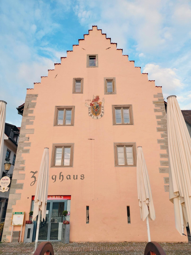 Rosa Gebäude mit einem zentralen Uhrenturm, umgeben von Fenstern, Regenschirmen, einem Schild, einer Straßenlaterne, Topfpflanzen und einem bewölkten Himmel in München, Deutschland.