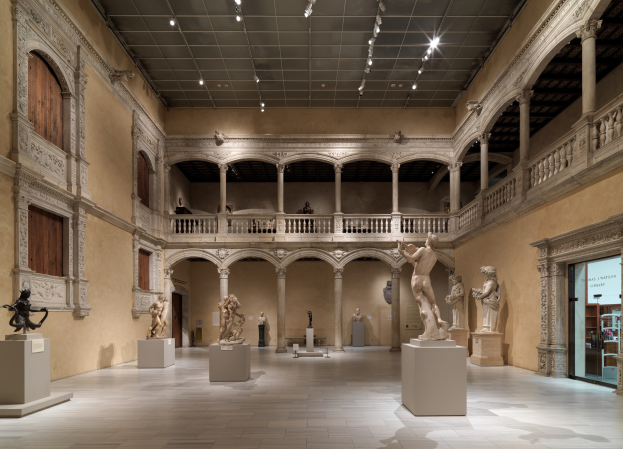 Ein geräumiger Museumsgalerie mit zahlreichen Statuen und Skulpturen auf Sockeln, mit architektonischen Elementen wie Wänden, Säulen, Geländern und einer beleuchteten Decke sowie einigen Besuchern.