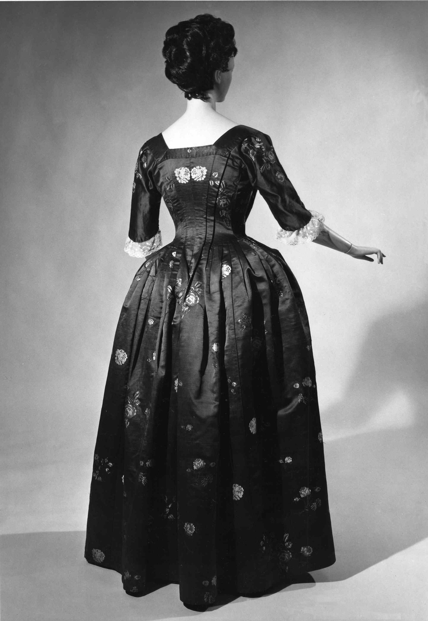 Eine Frau in einem Schwarz-Weiß-Foto aus den 1950er Jahren trägt ein langes, enges Kleid mit einem weiten Rock, hält ihre Hände zusammen und hat eine klassische Hochsteckfrisur mit dezentem Make-up und schwarzen Pumps.