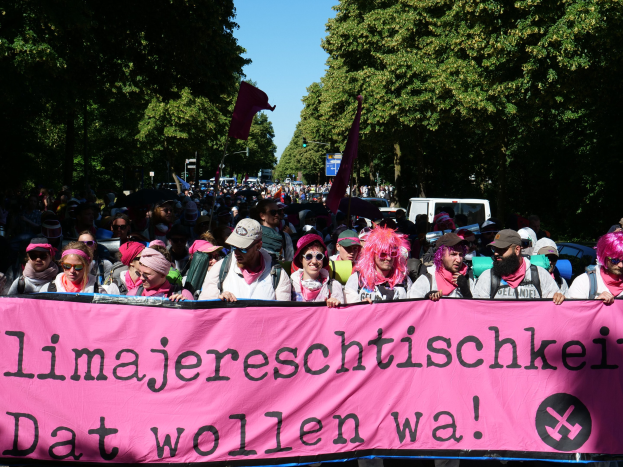 Eine Gruppe von Menschen marschiert mit einem pinken Banner, auf dem 'Frauenmarsch in Berlin' steht, die Straße entlang, einige tragen Mützen und Schutzbrillen, andere halten Fahnen, vor einem Hintergrund aus Bäumen, Fahrzeugen und Himmel.
