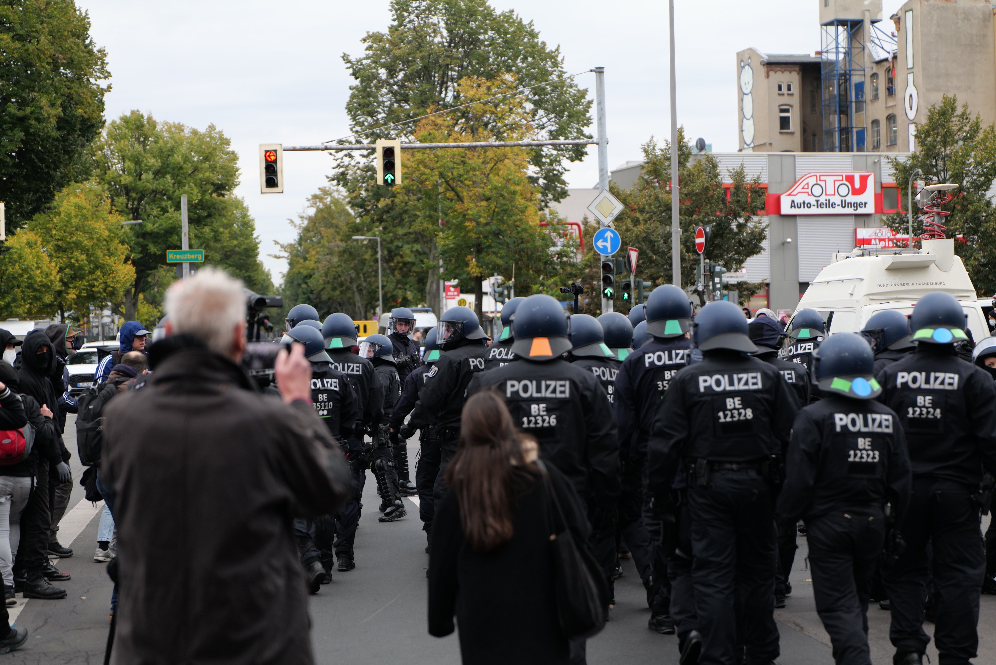 Eine Gruppe von Menschen, die eine Straße entlanggehen, mit Polizeibeamten, einige mit Helmen und Taschen oder Kameras, sowie Fahrzeugen, Verkehrszeichen, Schildern, Bäumen, Gebäuden und einem klaren Himmel im Hintergrund.