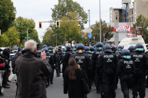 Eine Gruppe von Menschen, die eine Straße entlanggehen, mit Polizeibeamten, einige mit Helmen und Taschen oder Kameras, sowie Fahrzeugen, Verkehrszeichen, Schildern, Bäumen, Gebäuden und einem klaren Himmel im Hintergrund.
