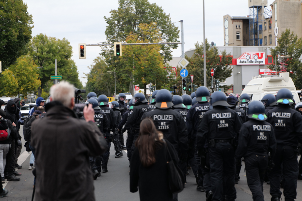 Eine Gruppe von Menschen, die eine Straße entlanggehen, mit Polizeibeamten, einige mit Helmen und Taschen oder Kameras, sowie Fahrzeugen, Verkehrszeichen, Schildern, Bäumen, Gebäuden und einem klaren Himmel im Hintergrund.
