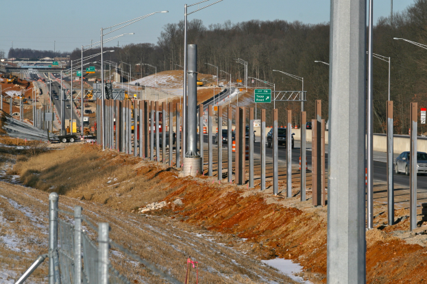 Baustelle mit Fahrzeugen, Pfählen, Lampen, Beschilderung, einem Zaun, schneebedecktem Gras, Bäumen und einem Himmel im Hintergrund.