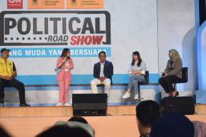 Eine Gruppe von Menschen sitzt auf einer Bühne mit einer Frau in der Mitte, die ein Mikrofon hält, ein Publikum im Vordergrund und ein Banner mit der Aufschrift "Politischer Straßenumzug 2019" im Hintergrund.