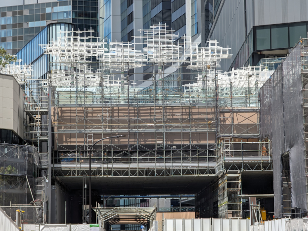 Großes Gebäude mit Gerüst, das in einer städtischen Umgebung von Bäumen links und einer Straße unten umgeben ist.
