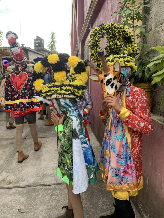 Eine Gruppe von Menschen in bunten Kostümen steht vor einem Gebäude während eines Faschingsumzugs, mit Bäumen, Gebäuden und einem klaren blauen Himmel im Hintergrund.