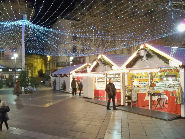 Ein belebter Weihnachtsmarkt bei Nacht in einer Stadt, mit Menschen, die stehen, sitzen und ein Kind halten, dekoriert mit festlichen Lichtern, Pflanzen und Gebäuden mit Fenstern unter einem bewölkten Himmel.