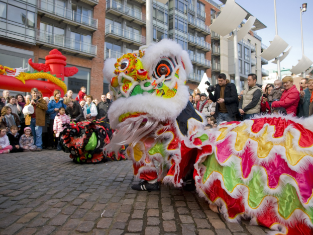 Ein lebendiges chinesisches Neujahrsfest in Amsterdam mit einem Drachentanz im Vordergrund und einer Menschenmenge, einige halten Kameras, vor einem Hintergrund aus Gebäuden, Laternenmästen und einem klaren blauen Himmel.
