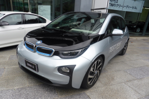 Ein weißer BMW i3 Elektroauto vor einem Gebäude in Tokyo, Japan, mit Glaswänden und einer Tafel im Hintergrund.