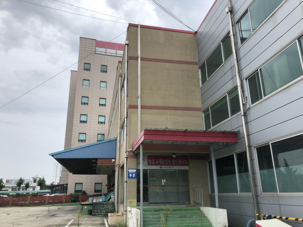 Großes modernes Gebäude mit vielen Fenstern, als Hauptquartier der Koreanischen Nationaluniversität für Wissenschaft und Technologie bezeichnet, umgeben von Grünflächen, einer Treppe, einem Schild, Pfählen, einem Zaun, Bäumen, Fahrzeugen, Stromleitungen und einem bewölkten Himmel.