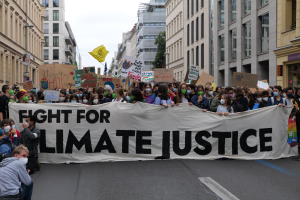 Eine Gruppe von Menschen marschiert mit einem Banner 'Kämpfe für Klimagerechtigkeit' und verschiedenen Plakaten die Straße entlang, trägt Masken, mit Gebäuden an der Straße und einem klaren blauen Himmel im Hintergrund.