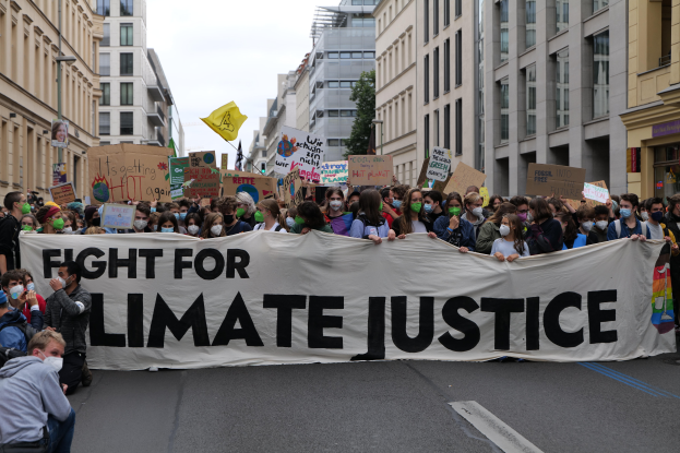 Eine Gruppe von Menschen marschiert mit einem Banner 'Kämpfe für Klimagerechtigkeit' und verschiedenen Plakaten die Straße entlang, trägt Masken, mit Gebäuden an der Straße und einem klaren blauen Himmel im Hintergrund.