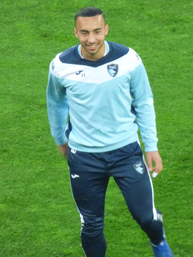 Ein lachender Mann in einem blauen Hemd und schwarzen Hosen läuft auf einem Fußballfeld, trägt sportliche Schuhe.