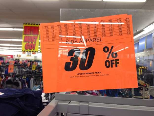 Ein Geschäft mit einem Schild "Kids Apparel 30% Off" im Vordergrund, Kleider im Hintergrund und eine Texttafel auf der rechten Seite.
