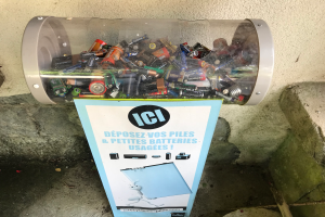 Ein Müllcontainer voller Batterien auf einem Gehweg, mit einem Plakat mit Text an der Wand dahinter und einer Treppe auf der rechten Seite.