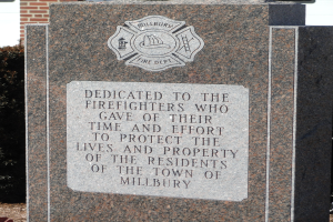 Denkmalstein mit einer Gedenktafel für Feuerwehrleute in Millbury vor einem Backsteingebäude mit umliegender Vegetation.