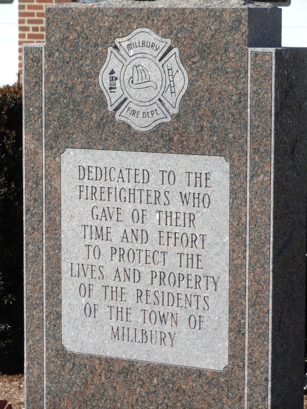 Denkmalstein mit einer Gedenktafel für Feuerwehrleute in Millbury vor einem Backsteingebäude mit umliegender Vegetation.