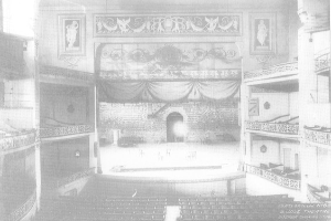Schwarzes und weißes Foto des Theatersaals des Theatre Royal London, das eine Bühne mit Stühlen, Geländern, skulptierten Wänden und einem Vorhang zeigt, mit Text am unteren Rand.