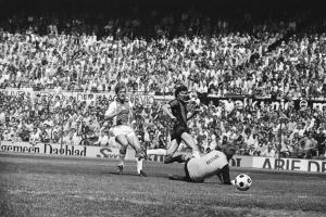 Schwarzes Foto von Männern, die auf einem Feld Fussball spielen, mit Zuschauern im Hintergrund und einer "Bundesliga 1978-79 Ajax 1-1 Hannover 96"-Tafel.