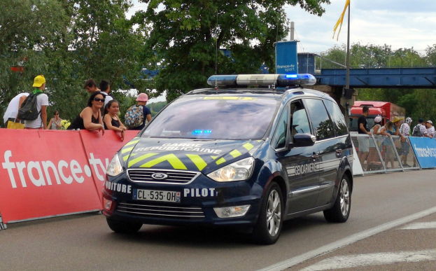 Polizeiauto fährt an einer Menschenmenge vorbei, die Schilder hölt, Bäume, eine Brücke, eine Flagge und einen bewölktem Himmel im Hintergrund.