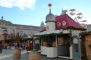 Ein geschäftiger Weihnachtsmarkt in Nürnberg, Deutschland mit Menschen um geschmückte Stände, einem Riesenrad im Hintergrund und einer Tafel mit Text auf der rechten Seite.