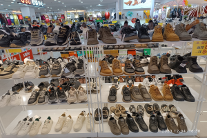 Ein Schuhgeschäftsinneres mit verschiedenen Schuhen auf Gestellen, Preisschildern, Menschen im Hintergrund, hängenden Kleidern, Deckenlampen, einem Bildschirm und Texttafeln, mit einem Wasserzeichen, das einen Laden in China anzeigt.