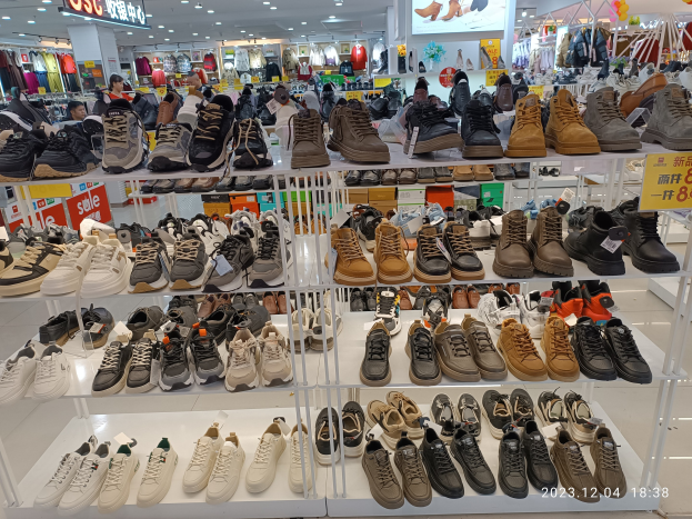 Ein Schuhgeschäftsinneres mit verschiedenen Schuhen auf Gestellen, Preisschildern, Menschen im Hintergrund, hängenden Kleidern, Deckenlampen, einem Bildschirm und Texttafeln, mit einem Wasserzeichen, das einen Laden in China anzeigt.
