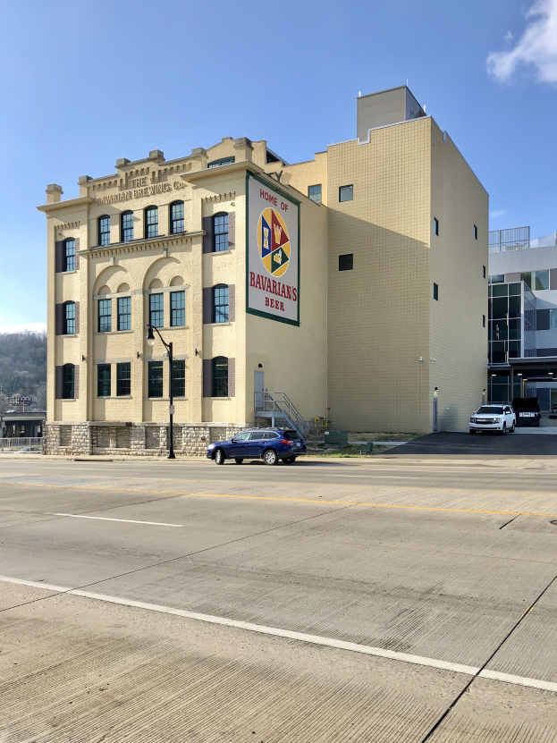 Großes Backsteingebäude mit "Home of Bavarian Brewing Co." -Schriftzug, Straßenlaternen, Fahrzeugen, Bäumen, Hügeln und bewölktem Himmel im Hintergrund.