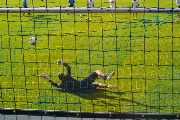 Ein Fußballtorhüter beim Abwehrversuch vor dem Tor, mit einem Netz im Vordergrund und einigen Menschen im Hintergrund, sowie mehreren Fußballen auf dem Boden verteilt.