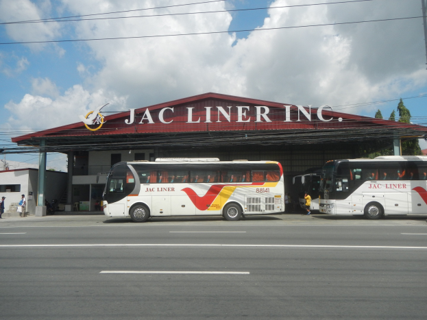 Zwei Busse vor einem Gebäude mit der Aufschrift "Jac Liner Inc." geparkt, Menschen auf dem Gehweg, Strommäste mit Drähten, Bäume und ein bewölkter Himmel im Hintergrund.