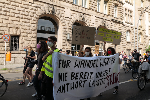 Eine Gruppe von Menschen marschiert auf der Straße in Berlin, hält Schilder und Banner hoch und fährt mit Fahrrädern, vor einem Gebäude mit Fenstern, Bögen, Säulen, Skulpturen, Bäumen und einem Laternenmast, protestiert gegen die Anti-Kriegsbewegung.