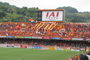 Fußballspiel in einem Stadion mit großer Zuschauermenge, saftig grünem Rasen, Tor, Bannern, Fahnen, einem großen Bildschirm, Bäumen und einem klaren blauen Himmel.