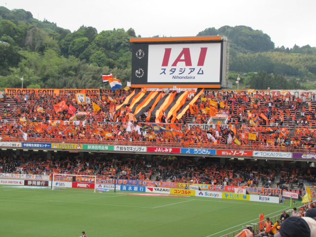Fußballspiel in einem Stadion mit großer Zuschauermenge, saftig grünem Rasen, Tor, Bannern, Fahnen, einem großen Bildschirm, Bäumen und einem klaren blauen Himmel.