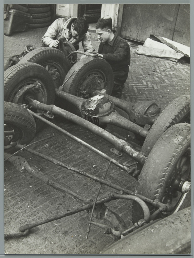 Zwei Männer in einer Garage, die Werkzeuge halten, arbeiten an einem Auto umgeben von Reifen und anderen Gegenständen, dargestellt in Schwarz-Weiß.