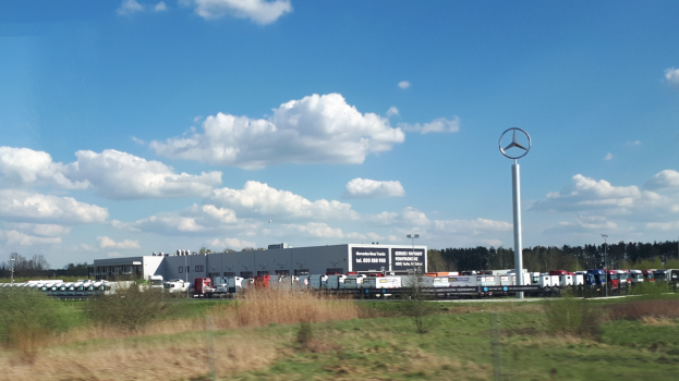 Ein Mercedes-Benz-Werk in Deutschland mit Fahrzeugen auf der Straße, umgeben von Gras, Pflanzen, Bäumen, Gebäuden und Polen, unter einem bewölkten Himmel.