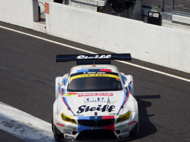 Ein BMW M6 GT3 Rennauto auf einer Rennstrecke mit einer Wand, einem Maschendrahtzaun und anderen Objekten im Hintergrund.