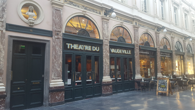 Theater du Vaudeville-Gebäude in Paris mit Türen, Fenstern, beleuchteter Beschilderung, Tischen, Stühlen und einer Wandstatue.