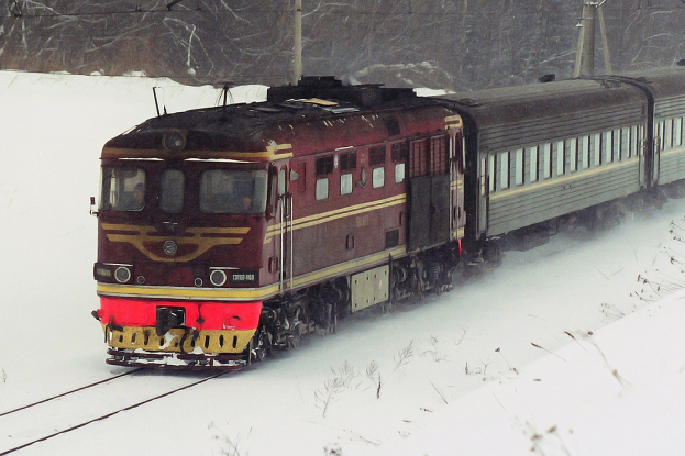 Ein Zug fährt auf schneebedeckten Schienen, mit Strommasten und Bäumen im Hintergrund, umgeben von einer Schneedecke.