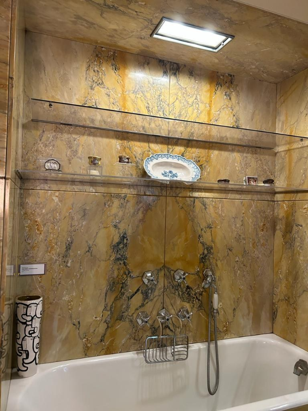 Bad mit Wanne und Dusche, mit Armatur, Ständer, Vase und Gegenständen auf Regalen, mit Marmorfliesen an den Wänden und beleuchteter Decke.
