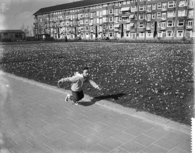 Ein junger Junge läuft über einen Gehweg in einem Park, umgeben von Gebäuden, Bäumen und einem klaren Himmel, und trägt Schuhe.