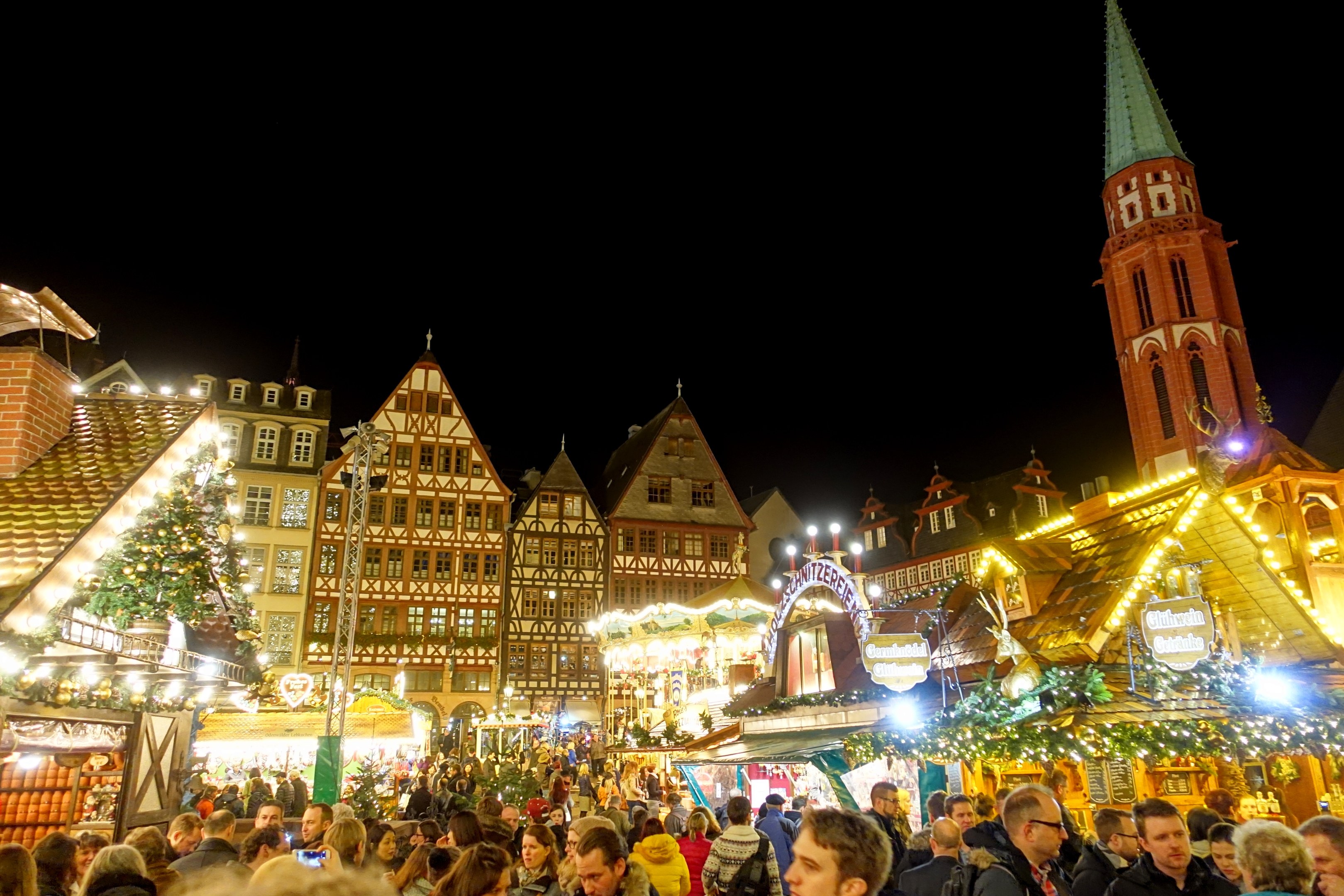 Ein geschäftiger Weihnachtsmarkt in Nürnberg, Deutschland mit Menschen um geschmückte Stände, festlicher Beleuchtung, Gebäude mit Fenstern, einem Uhrenturm und einem dunklen Himmel.