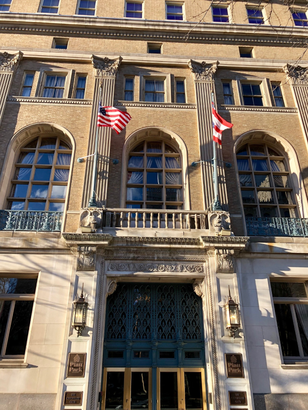 Großes Gebäude mit Glasfenstern, -türen, -säulen, -geländern, -leuchten, Schildern und Flaggen, als Plaza Hotel in New York City gegen einen Himmelhintergrund bezeichnet.
