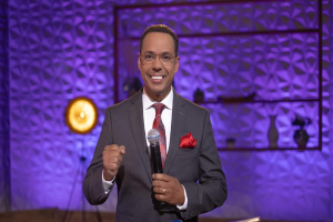 Stephen Colbert, der in einem Anzug, einer Krawatte und einer Brille auf einer Bühne steht und ein Mikrofon hält, mit einer Wand und verschiedenen Gegenständen dahinter.