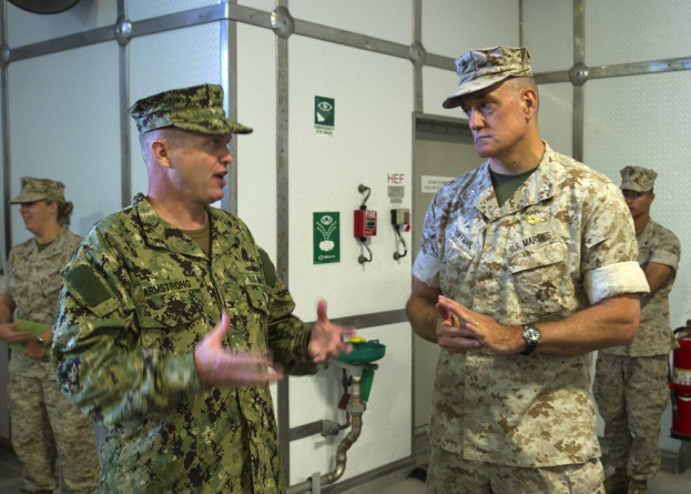 Zwei Männer in Militäruniformen unterhalten sich vor einer Wand mit einem Feuerlöscher auf der rechten Seite und Rohren und anderen Gegenständen im Hintergrund.