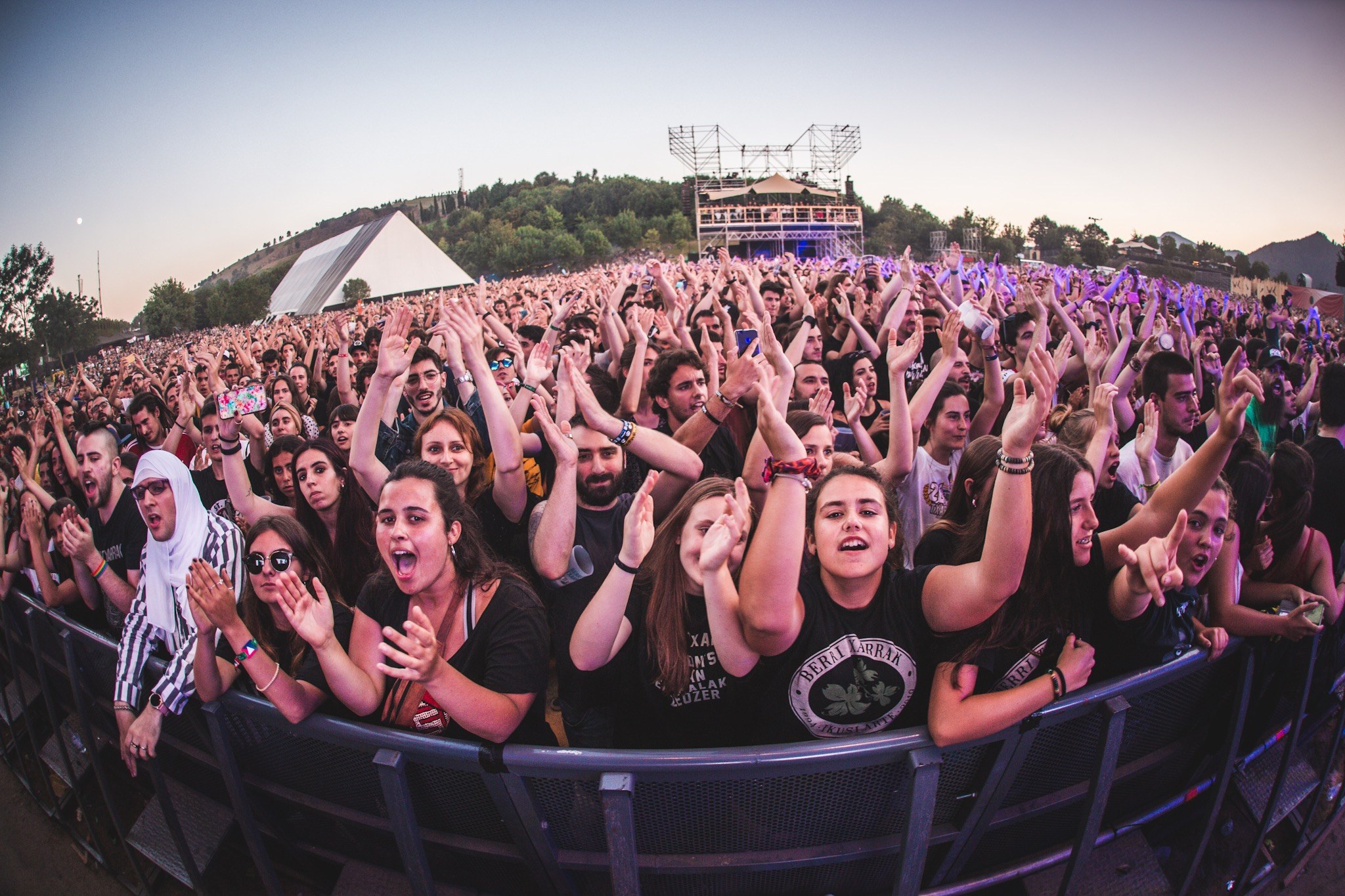 Eine große Menge bei einem Musikfestival mit erhobenen Händen vor einem Geländer, Bäumen, Gebäuden und einem klaren blauen Himmel im Hintergrund.
