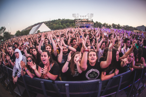 Eine große Menge bei einem Musikfestival mit erhobenen Händen vor einem Geländer, Bäumen, Gebäuden und einem klaren blauen Himmel im Hintergrund.