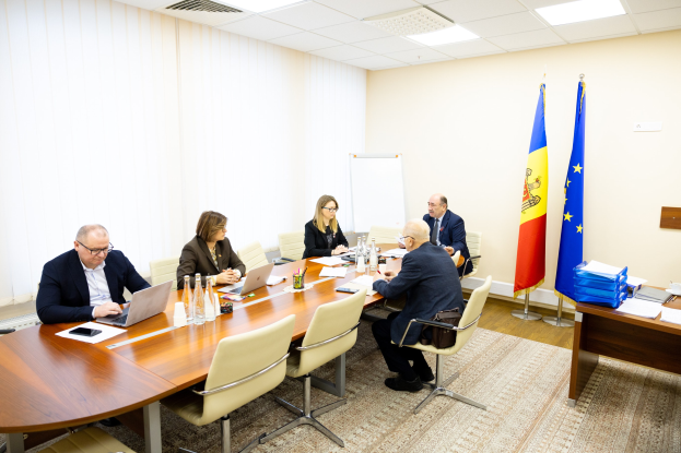 Eine Gruppe von Menschen sitzt um einen Konferenztisch mit Laptops, Flaschen, Gläsern, Papieren, Büchern und anderen Gegenständen und ist in eine Diskussion vertieft. Im Hintergrund sind Flaggen, eine Whiteboard und Deckenleuchten zu sehen.