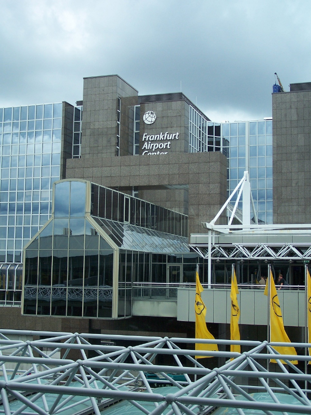 Ein großes Gebäude mit Glaswänden und Text, umgeben von gelben Flaggen und Eisenstangen, unter einem Himmel voller weißer, flauschiger Wolken, das den Frankfurter Flughafen in Frankfurt, Deutschland, zeigt.