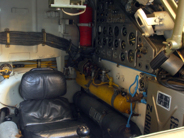 Cockpit einer kleinen Maschine mit schwarzem Ledersitz, verschiedenen Messgeräten, Feuerlöscher und weiterer Ausrüstung.
